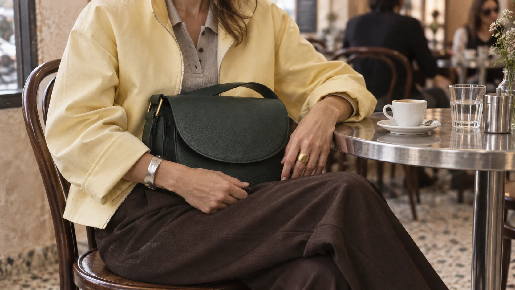 Una mujer con chaqueta amarilla sostiene un bolso verde sentada en un café.